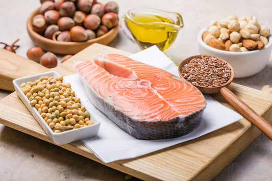 Eine Scheibe Lachs liegt auf einem Holzbrett, darunter ein Küchenpapier und daneben noch ein Holzlöffel mit Samen und eine rechteckige Schale mit Kichererbsen. Hinter dem Brett stehen noch zwei Nussschalen und eine kleine Karaffe mit Öl.