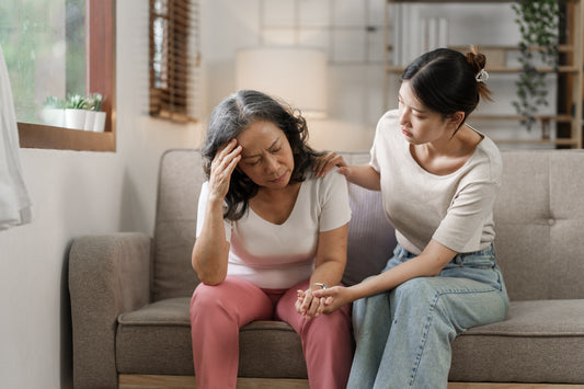 Auf einer beigen couich sitzen zwei Frauen. Die ältere Dame links sitzt vorgebeugt und hält sich die Hand an den Kopf. Die andere rechts hält ihre Hand und die andere Hand liegt auf ihrer Schulter. 