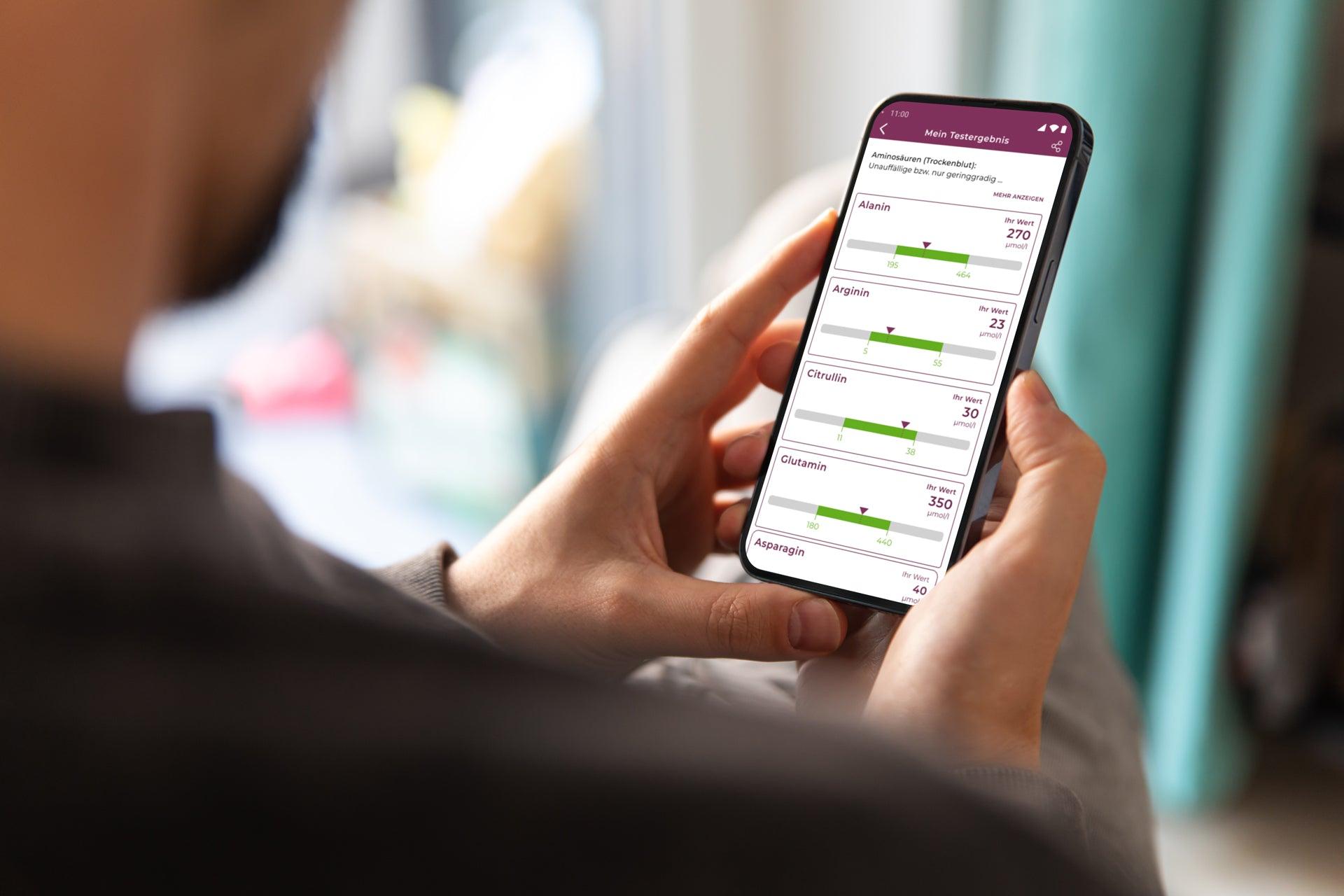 Über Schulterblick: Mann hält sein Telefon in der Hand. Wir sehen den Bildschirm mit cem Testergebnis drauf. Bei dem Testergebnis des Aminosäuren-Profils sieht man die Werte Alanin, Arginin, Citrullin und Glutamin. Die Werte werden in einer Skala angegeben. 